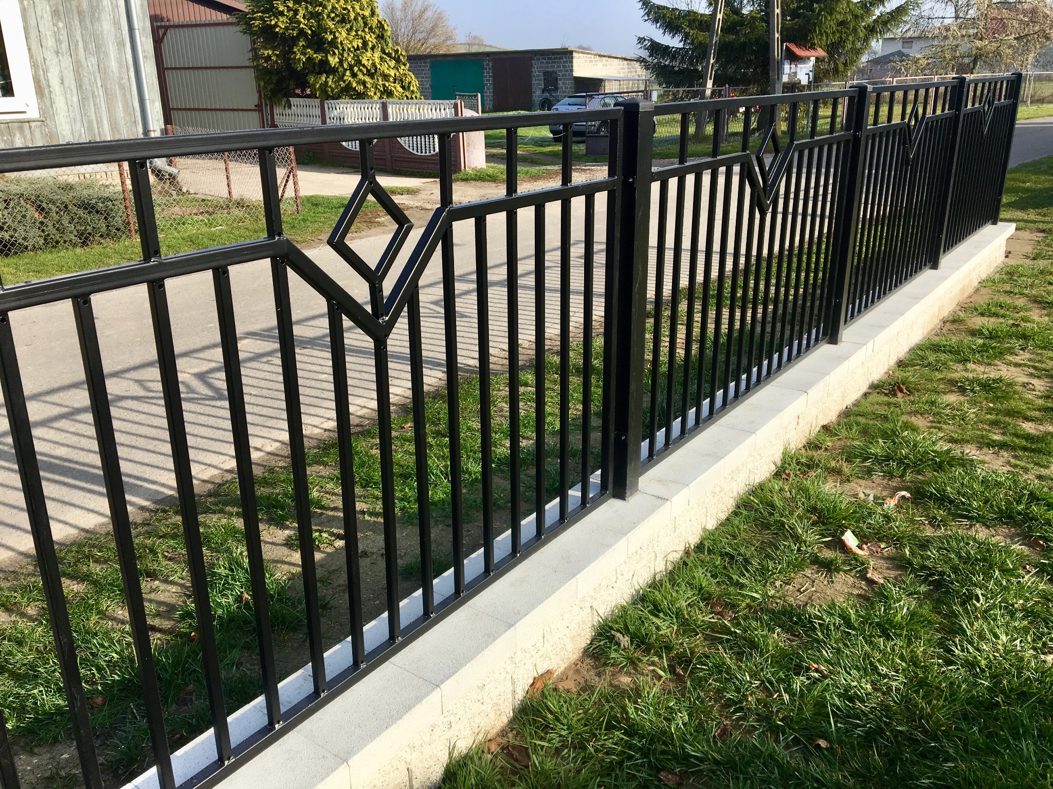 Czarny metalowy płot z geometrycznym wzorem diamentu osadzony na niskim betonowym murku, widok z boku na tle trawnika i zabudowań.