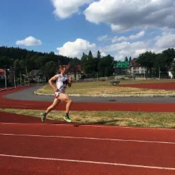 Mężczyzna w trakcie biegu na czerwonej bieżni stadionu, z zielonymi butami i numerem na koszulce, na tle drzew i błękitnego nieba z chmurami.