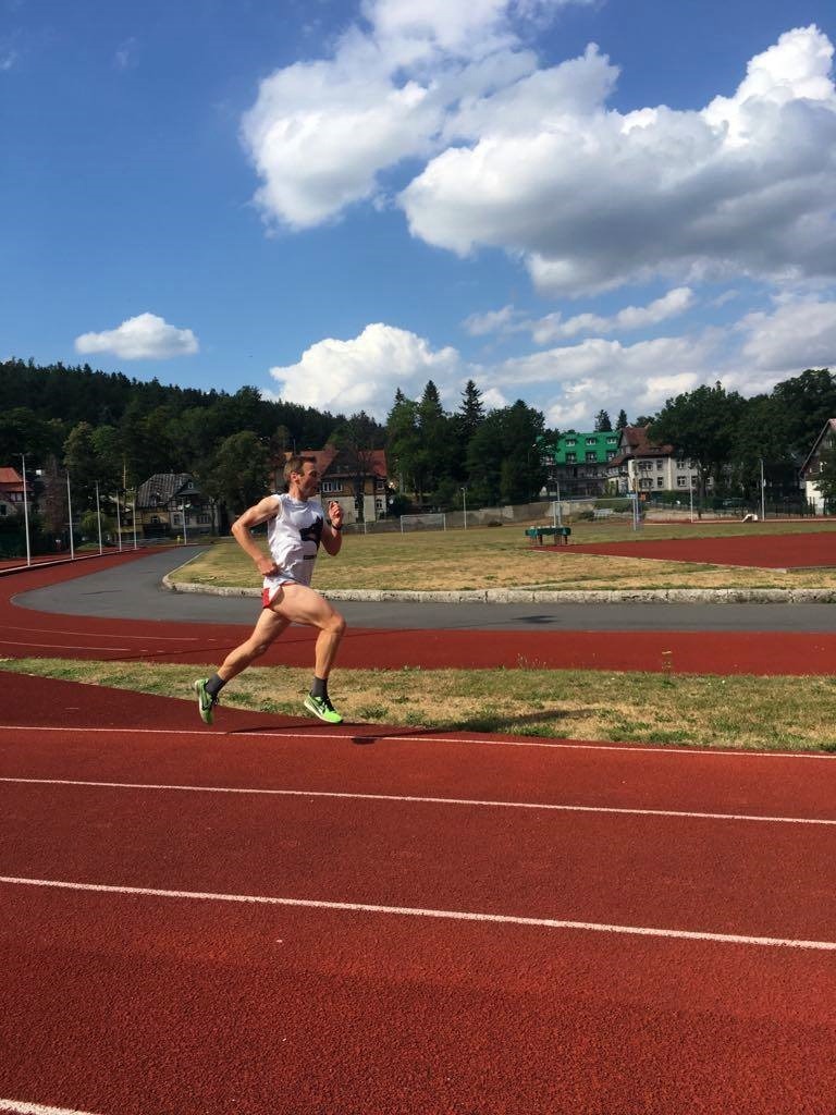 Mężczyzna w trakcie biegu na czerwonej bieżni stadionu, z zielonymi butami i numerem na koszulce, na tle drzew i błękitnego nieba z chmurami.