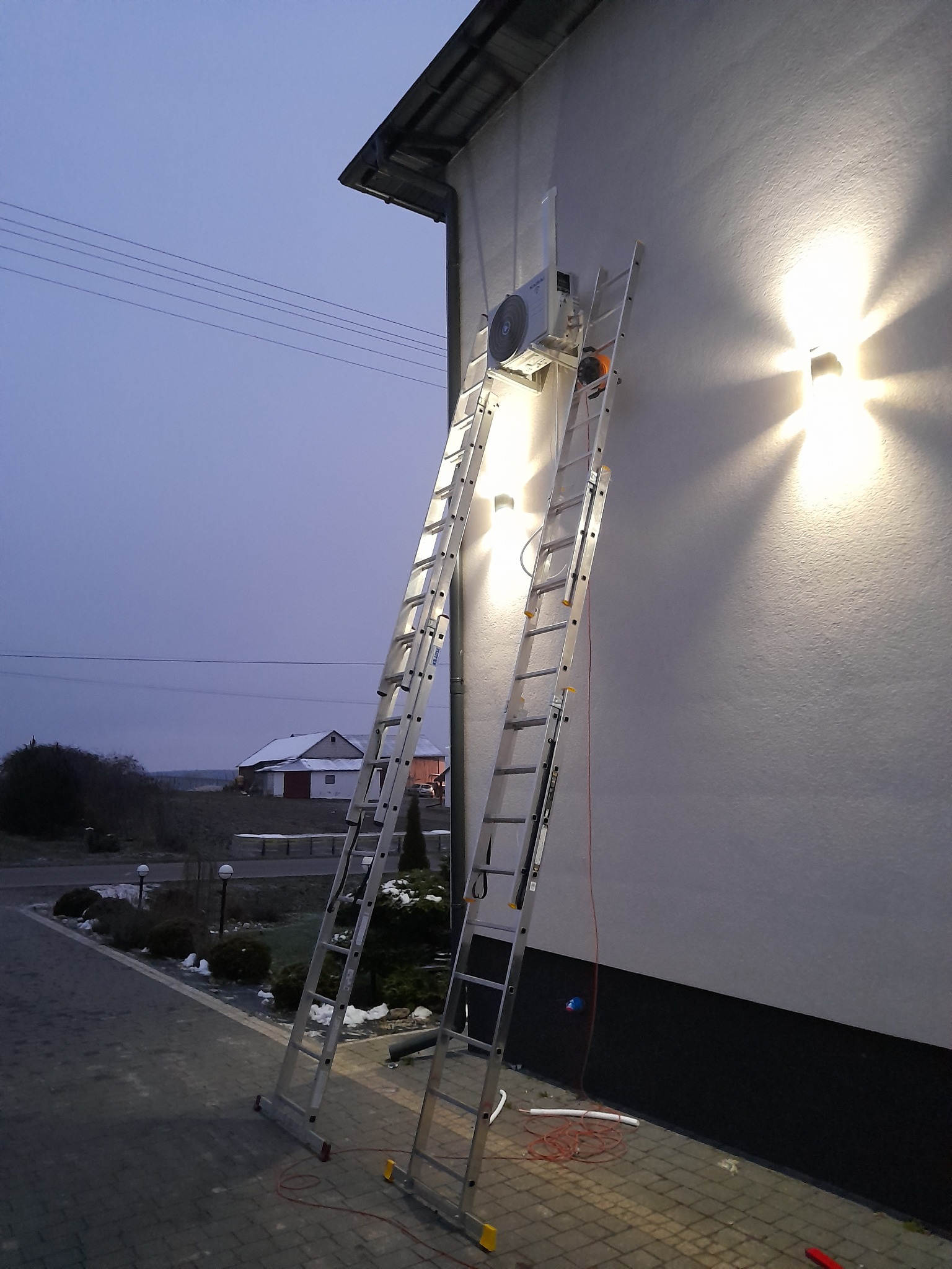 Zewnętrzna jednostka klimatyzacji zamontowana na ścianie domu, widoczna z poziomu chodnika, z rozstawioną drabiną aluminiową i przewodami na ziemi, oświetlona lampą elewacyjną.