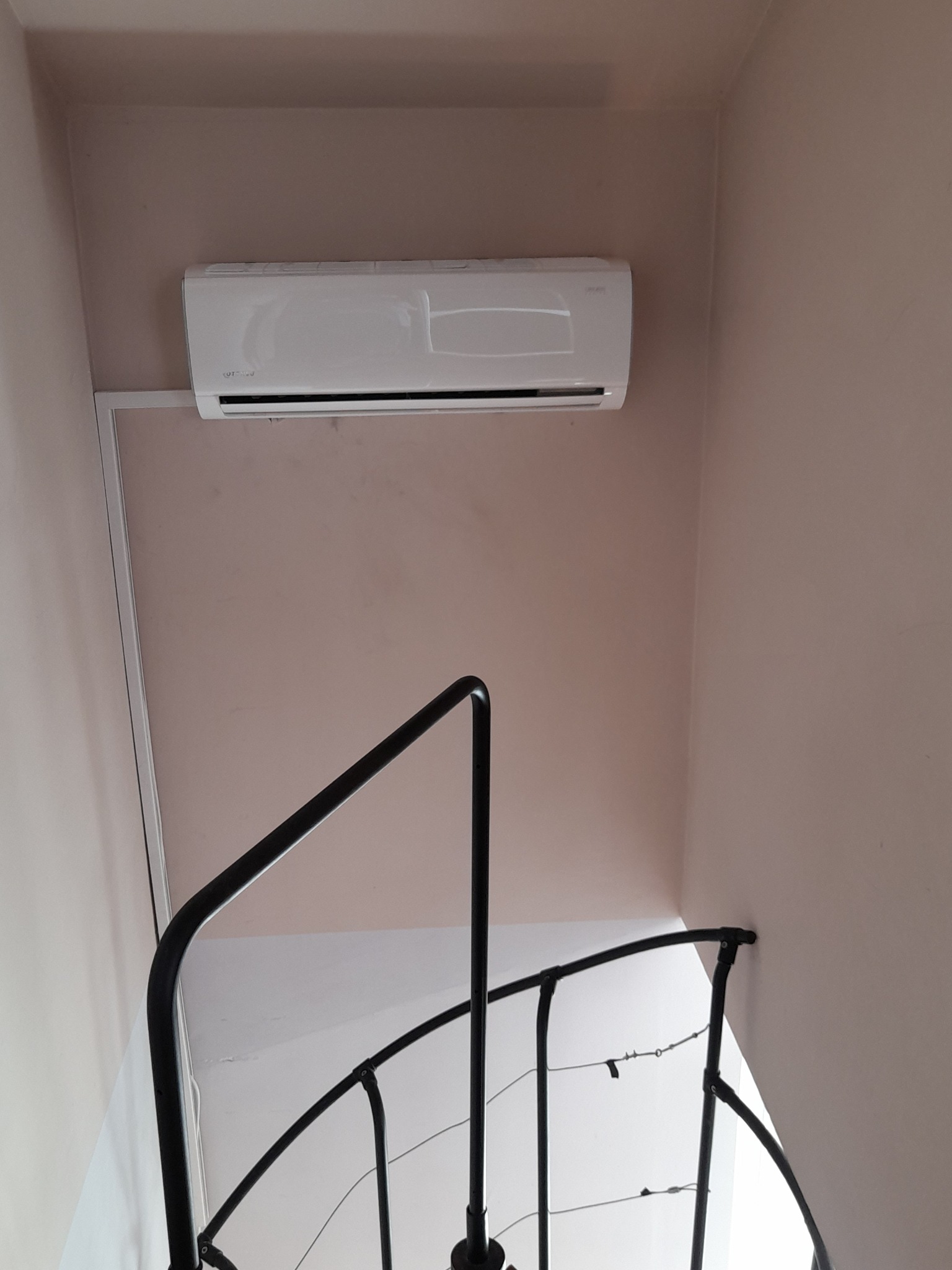 Biała jednostka klimatyzacji ściennej zamontowana w rogu pokoju o różowych ścianach, widoczna z perspektywy dolnej, z fragmentem czarnej, kręconej balustrady schodów w kadrze.