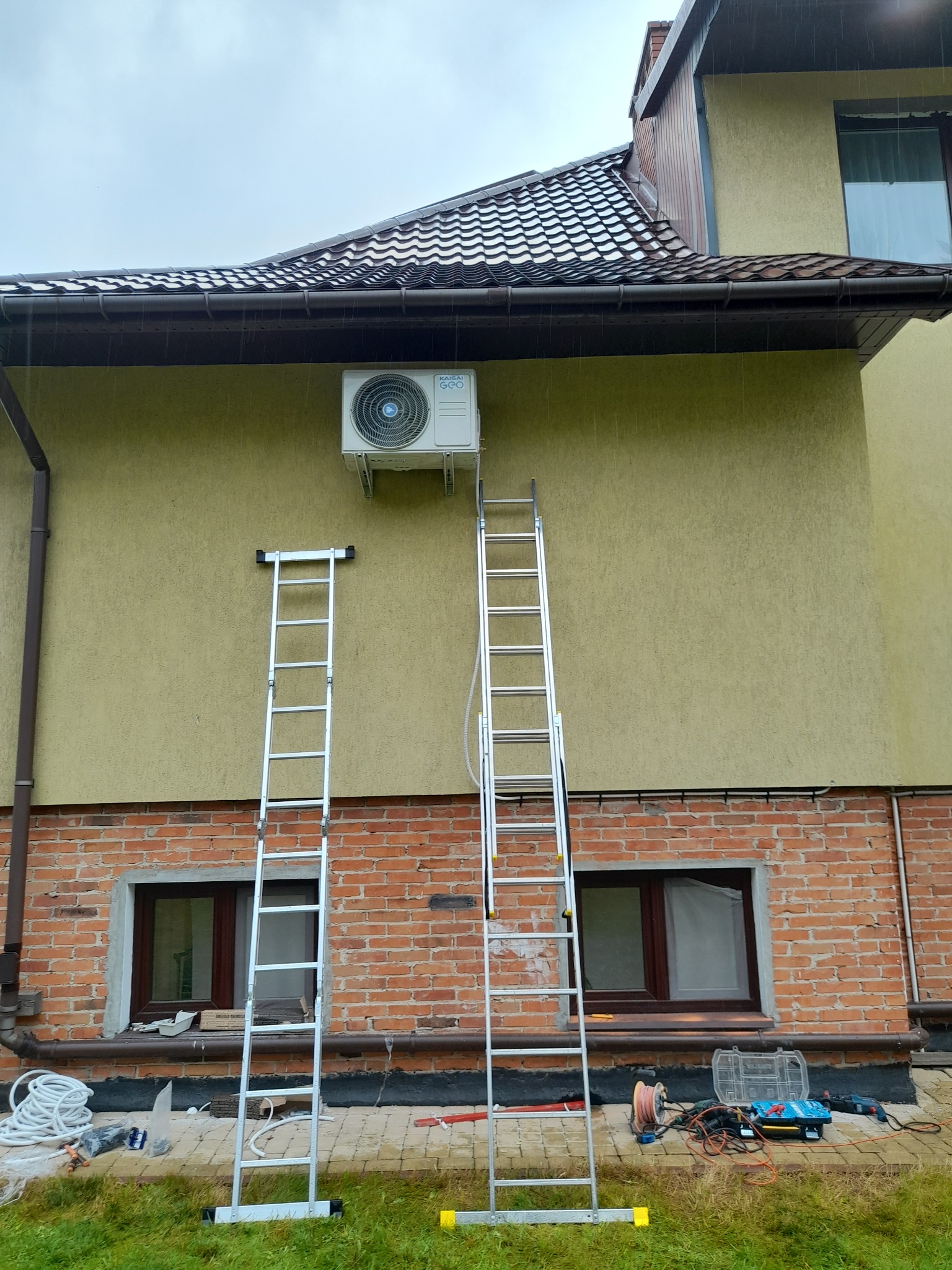 Zewnętrzna jednostka klimatyzatora zamontowana na elewacji domu, dwie drabiny opierają się o ścianę, widoczne narzędzia i przewody na trawie, instalacja w trakcie deszczu.
