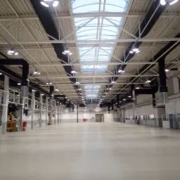 Wnętrze hali przemysłowej z widocznymi lampami sufitowymi, instalacją wentylacyjną i pracownikiem na podnośniku koszowym w oddali, przygotowującym się do pracy na wysokości.