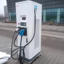Biała stacja ładowania pojazdów elektrycznych ABB z trzema złączami, umieszczona na kostce brukowej przed nowoczesnym budynkiem biurowym w pochmurny dzień. W tle widoczny pracownik w pomarańczowej...