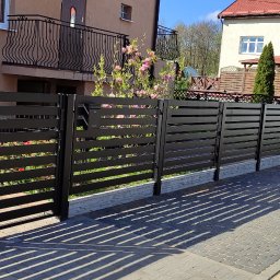 METALSTYL Mateusz Borek - Nowoczesne, poziome, czarne ogrodzenie panelowe z furtką i skrzynką na listy, z widocznym fragmentem elewacji domu z balkonem z kutą balustradą oraz zaparkowanym busem w tle.