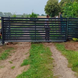 METALSTYL Mateusz Borek - Montaż przesuwnej bramy wjazdowej z poziomych profili stalowych, widoczne fundamenty i mechanizm jezdny, teren po pracach ziemnych, w tle łąka z balotami siana.