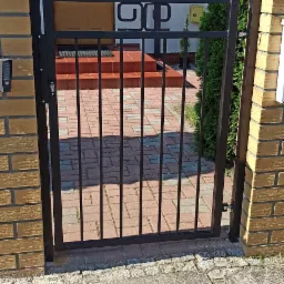 Czarna furtka z metalowymi prętami i ozdobnym elementem w górnej części, zamontowana między murowanymi słupkami z cegły klinkierowej, prowadząca do domu z drewnianymi drzwiami i schodami wyłożonymi...