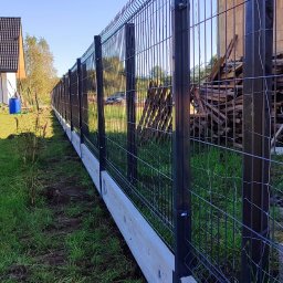 METALSTYL Mateusz Borek - Nowoczesne ogrodzenie panelowe z podmurówką betonową na tle domu jednorodzinnego i zielonego trawnika z piłką. Widoczne elementy konstrukcyjne i materiały budowlane.