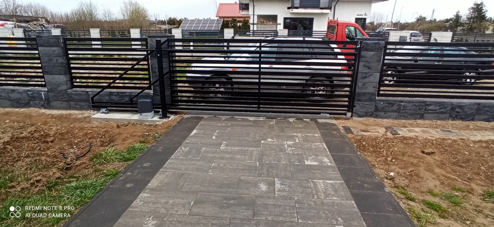 Automatyczna brama wjazdowa z poziomych profili stalowych, zamontowana między słupkami z szarej kostki łupanej, z widocznym napędem elektrycznym i podjazdem z szarej kostki brukowej.