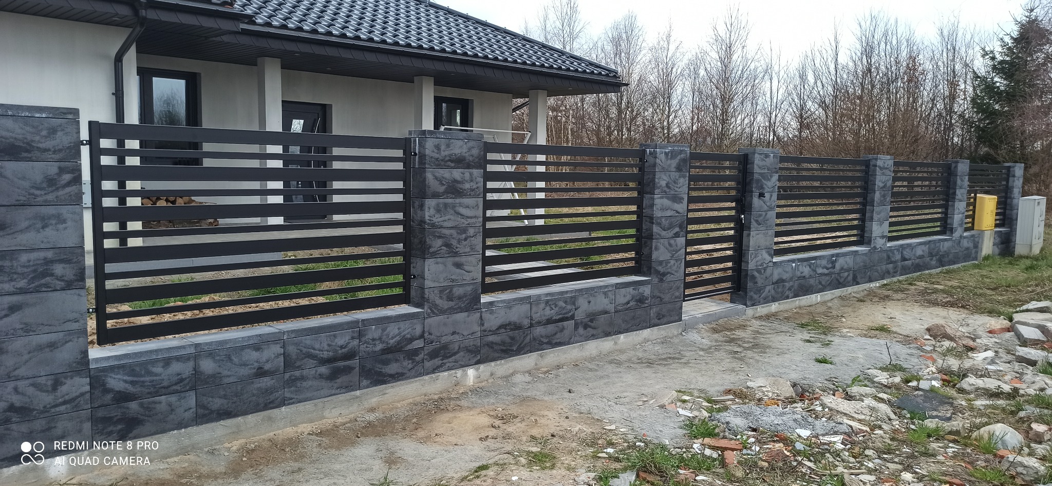 Nowoczesne ogrodzenie z poziomych, czarnych paneli metalowych i szarych, kamiennych słupków, widok na dom z ciemnymi oknami i stertą drewna za ogrodzeniem.