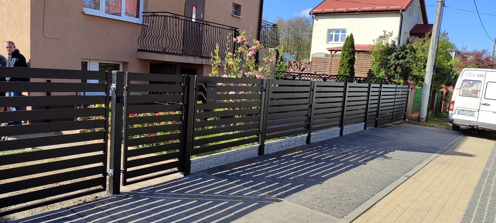 Nowoczesne, poziome, czarne ogrodzenie panelowe z furtką i skrzynką na listy, z widocznym fragmentem elewacji domu z balkonem z kutą balustradą oraz zaparkowanym busem w tle.