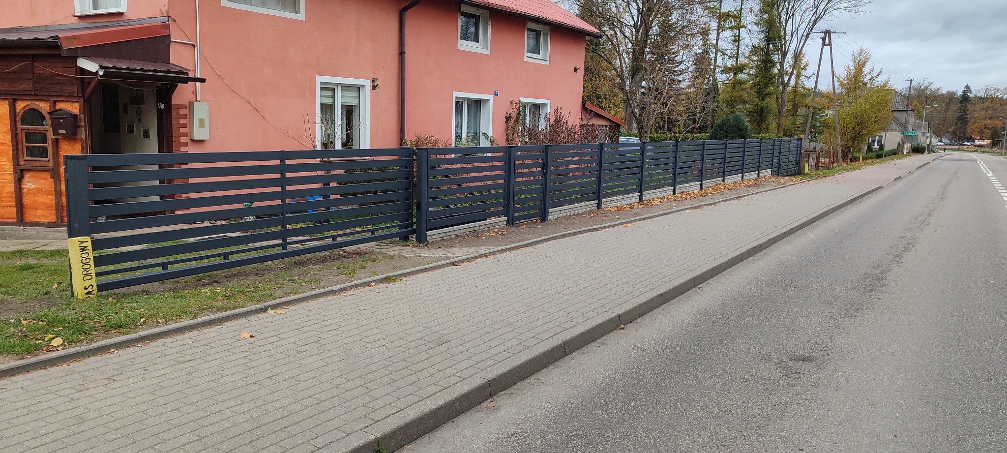 Nowoczesne, poziome ogrodzenie panelowe w kolorze grafitowym, oddzielające posesję z różowym domem od chodnika i ulicy, widoczne z perspektywy ulicy.