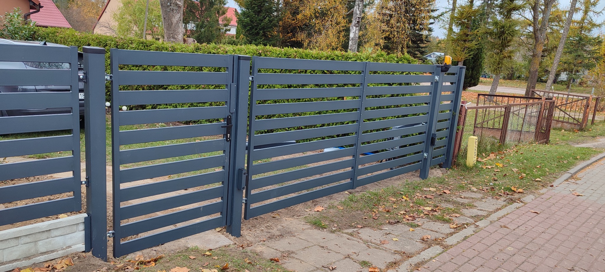 Nowoczesna, grafitowa brama wjazdowa z poziomymi panelami, zamontowana na posesji z zielonym żywopłotem w tle, widoczny automat do bramy i fragment samochodu za bramą.