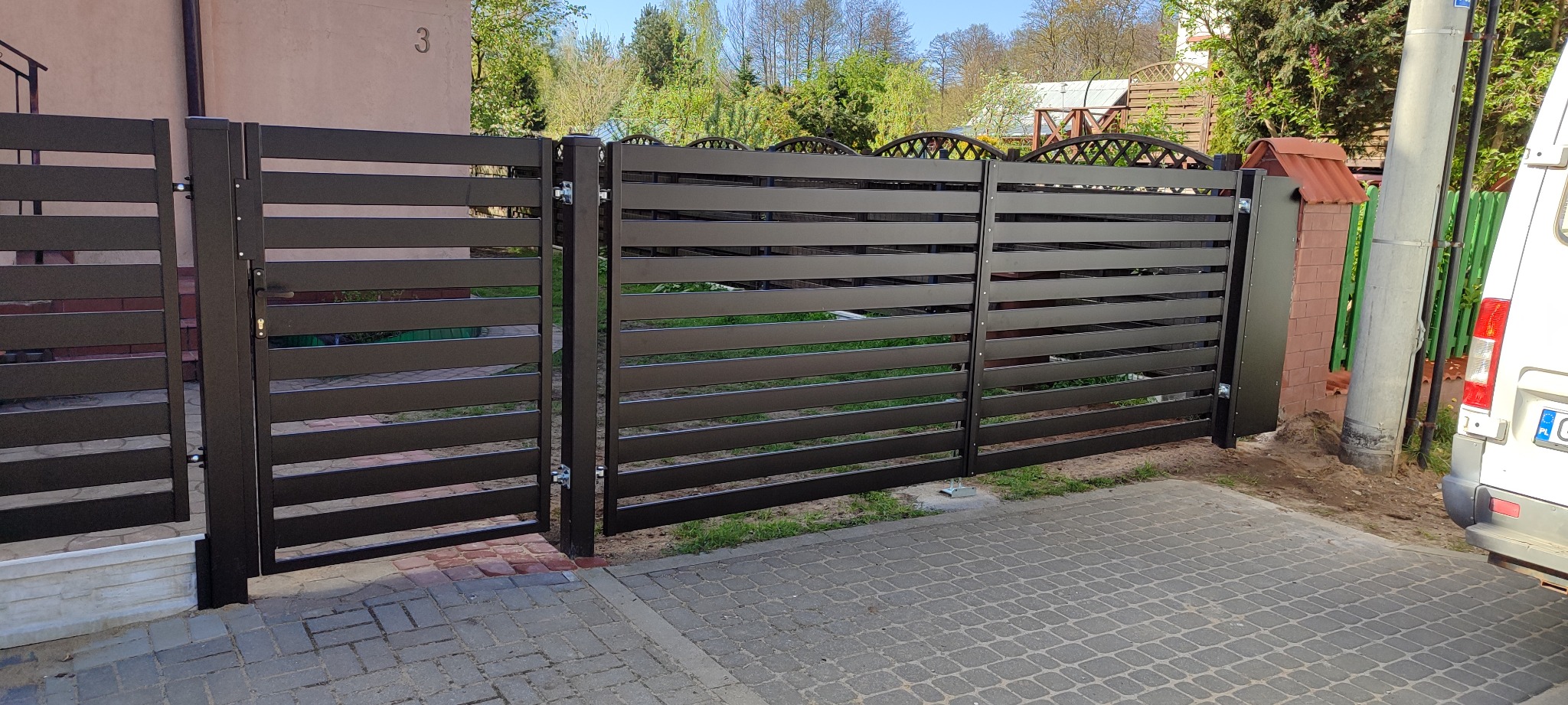 Nowoczesna, pozioma brama wjazdowa i przęsła ogrodzeniowe w kolorze grafitowym, z widocznymi elementami montażowymi, na tle kostki brukowej i zieleni.