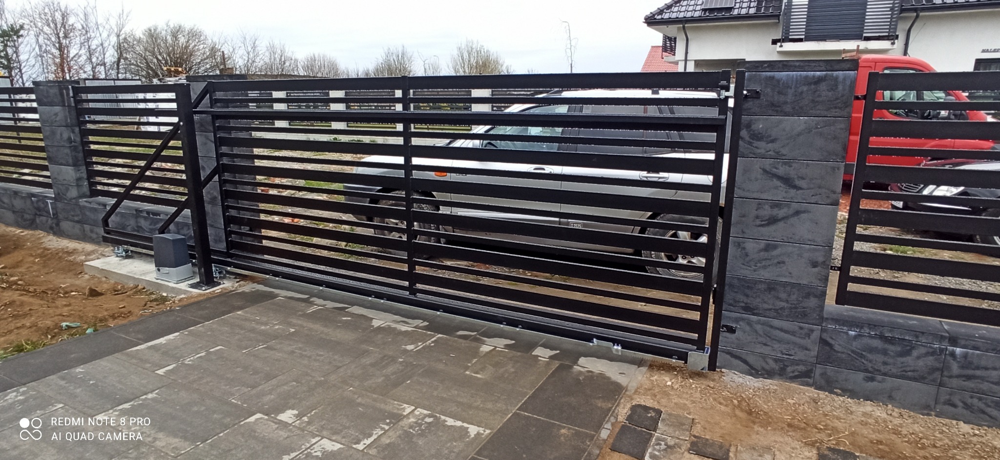 Nowoczesna, przesuwna brama wjazdowa z poziomymi, czarnymi profilami stalowymi, zamontowana na słupkach z szarej, imitującej kamień kostki, widoczny automat do bramy, na podjeździe szara kostka...
