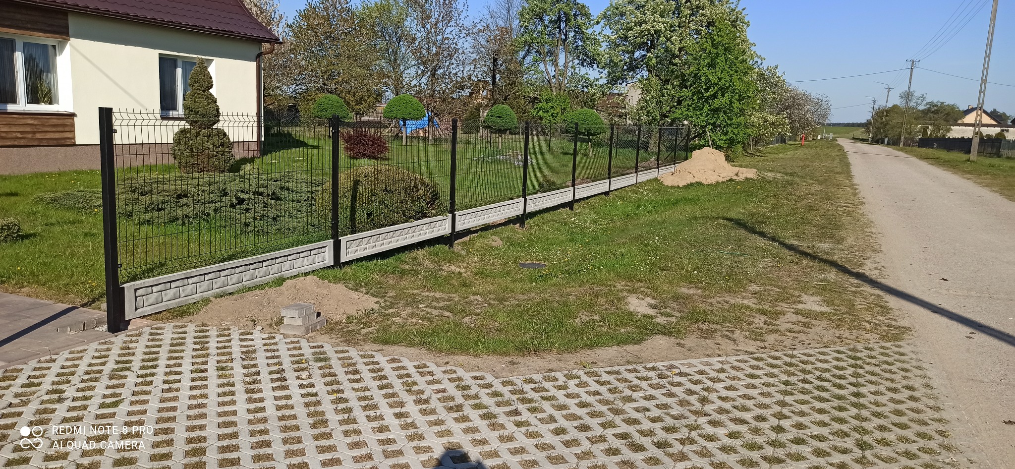 Nowoczesne ogrodzenie panelowe z podmurówką betonową o wzorze cegiełek, widok z perspektywy niskiego kąta na posesję z zadbanym trawnikiem i ozdobnymi krzewami.