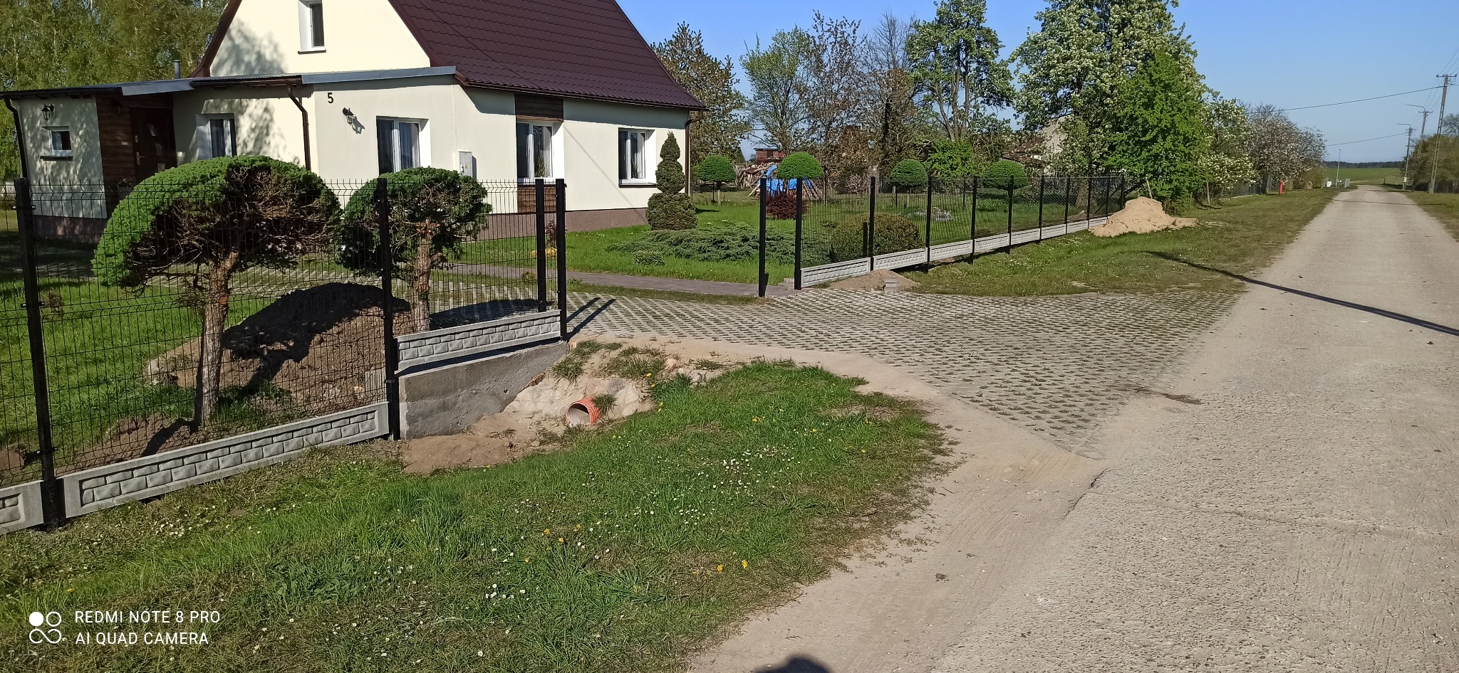 Ogrodzenie panelowe z podmurówką betonową i furtką, widok na dom z brązowym dachem, kostka ażurowa na podjeździe, uformowane krzewy.