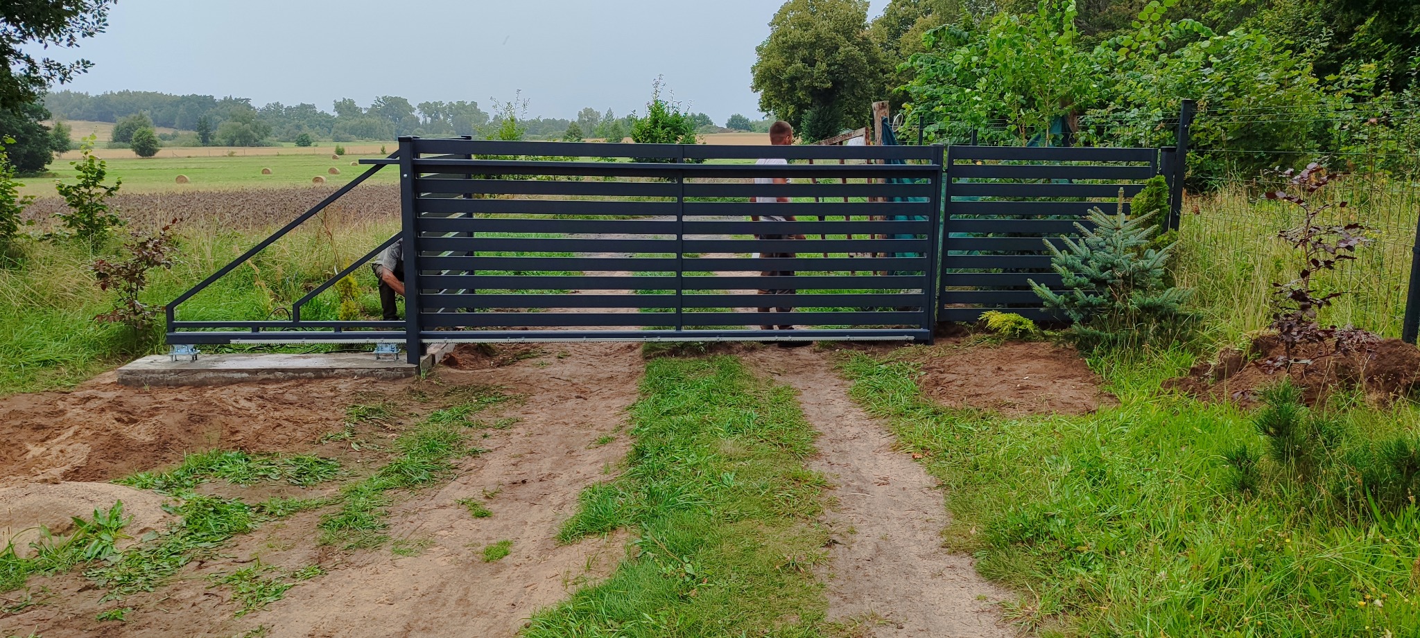 Montaż przesuwnej bramy wjazdowej z poziomych profili stalowych, widoczne fundamenty i mechanizm jezdny, teren po pracach ziemnych, w tle łąka z balotami siana.