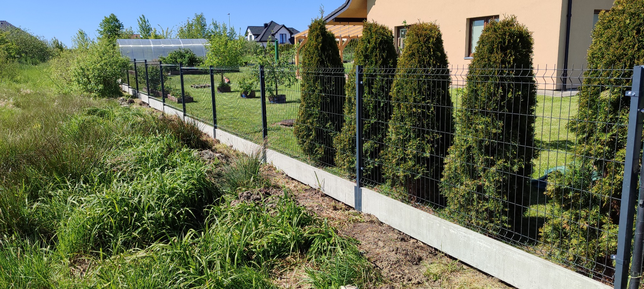 Nowoczesne ogrodzenie panelowe z podmurówką betonową, widok z zewnątrz posesji, z tujami posadzonymi wzdłuż ogrodzenia.