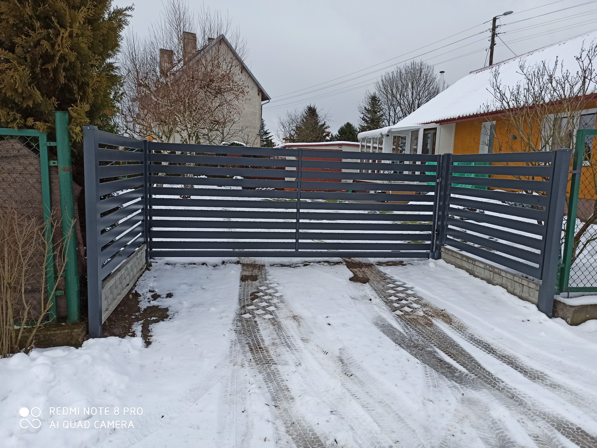 Nowoczesna, pozioma, szara brama wjazdowa z metalowych paneli na tle zaśnieżonego podjazdu i zabudowań mieszkalnych w pochmurny dzień.