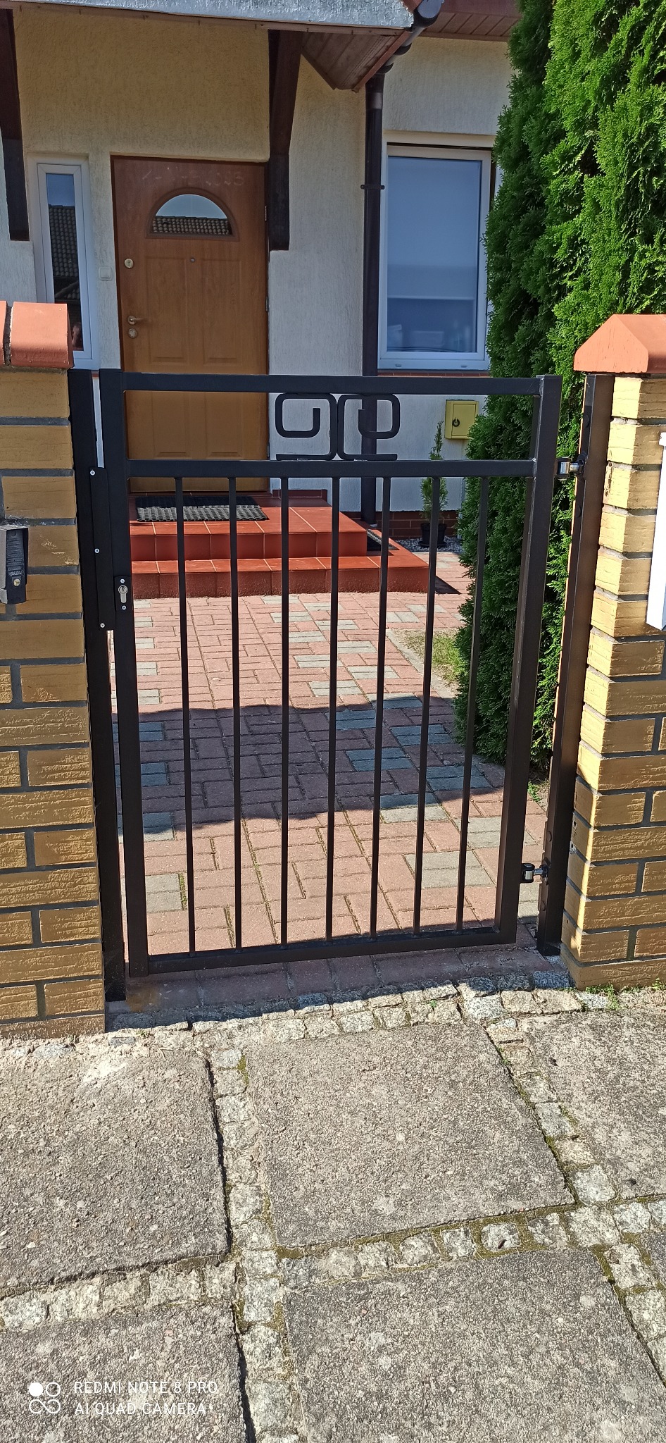 Czarna furtka z metalowymi prętami i ozdobnym elementem w górnej części, zamontowana między murowanymi słupkami z cegły klinkierowej, prowadząca do domu z drewnianymi drzwiami i schodami wyłożonymi...