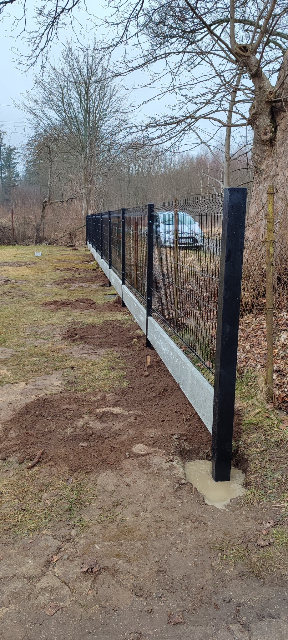 Montaż czarnego ogrodzenia panelowego z betonową podmurówką na terenie posesji, widoczne słupki zalane betonem, w tle zaparkowany samochód.