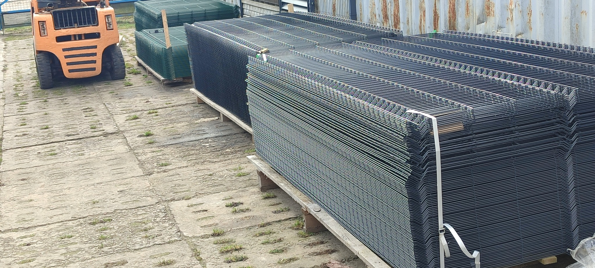 Stosy paneli ogrodzeniowych 3D w kolorze grafitowym i zielonym, ułożone na paletach, z pomarańczowym wózkiem widłowym w tle.