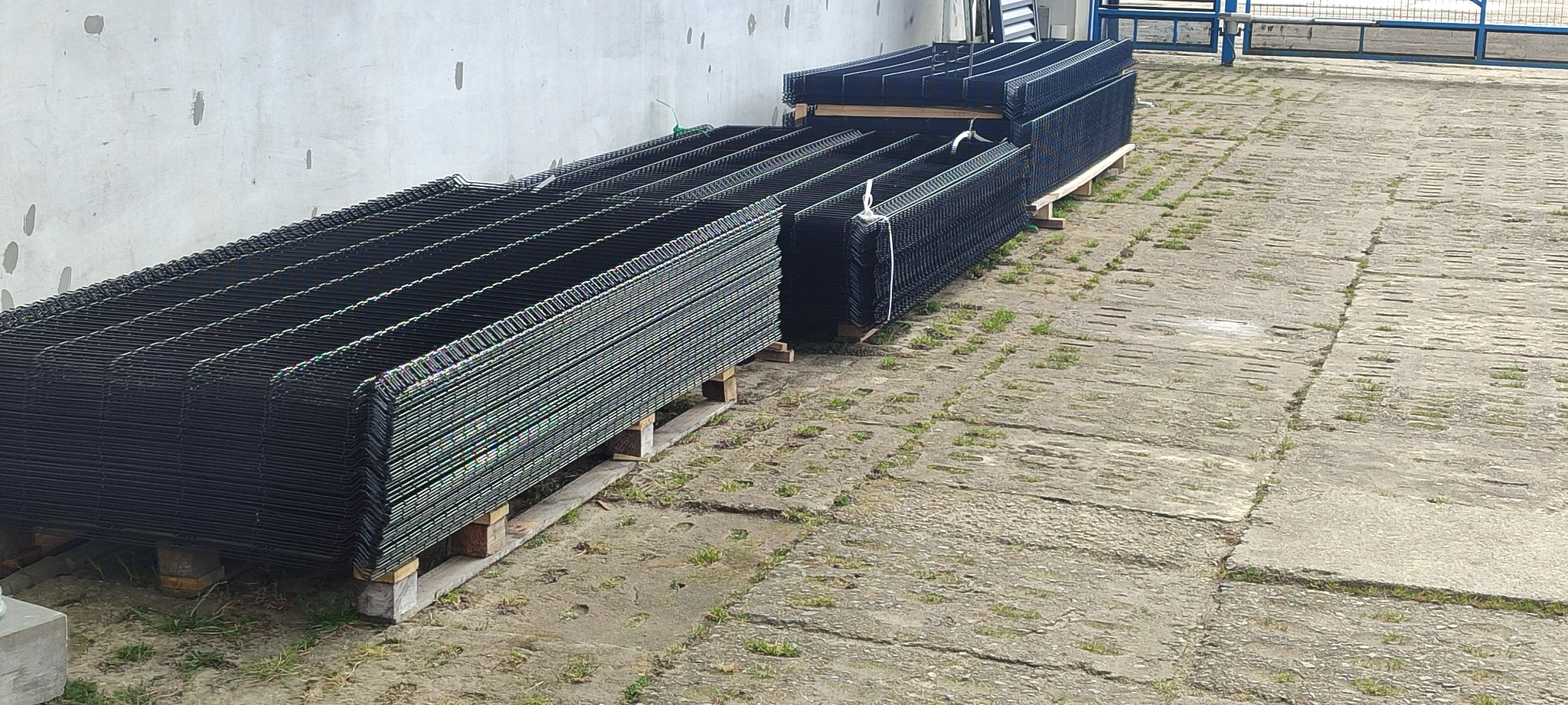 Stosy czarnych, metalowych paneli ogrodzeniowych ułożone na drewnianych paletach na betonowym placu z widoczną ścianą budynku w tle.