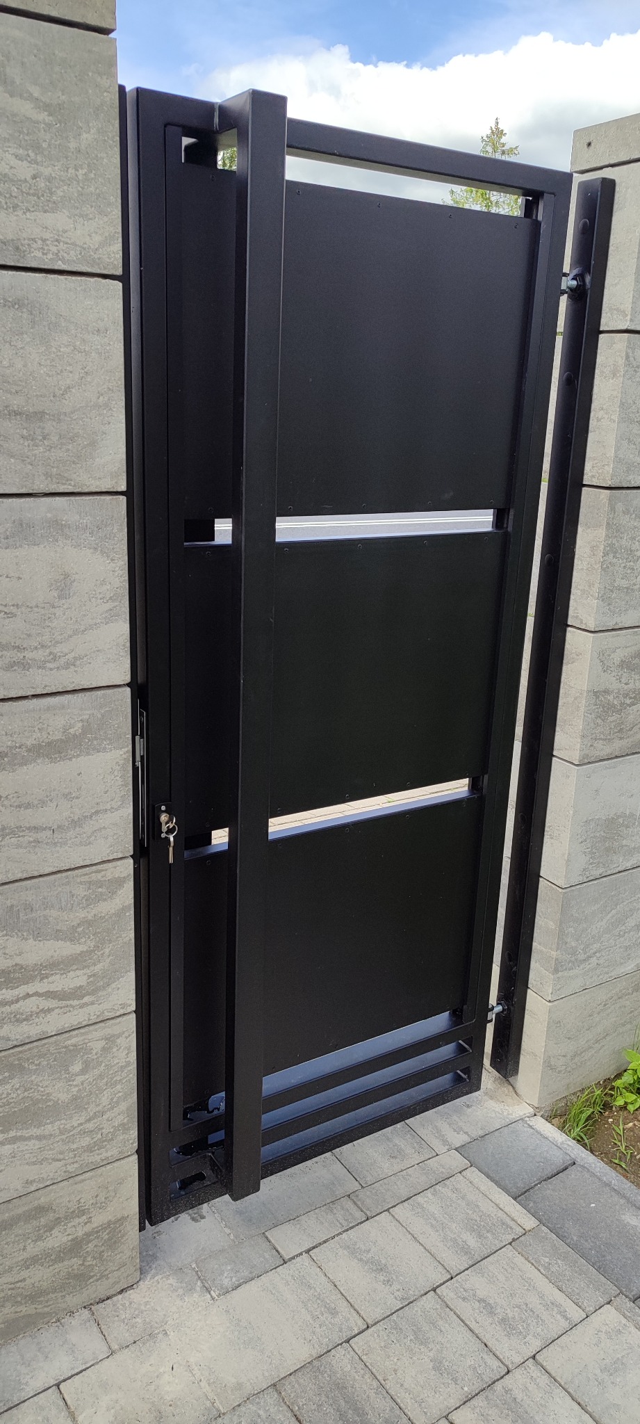 Nowoczesna, czarna brama wjazdowa z poziomymi prześwitami, zamontowana między betonowymi słupkami. Widoczne klucze w zamku. Minimalistyczny design.