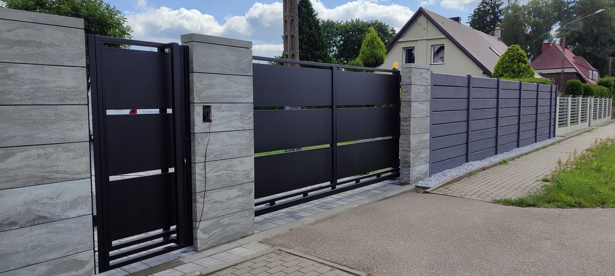 Nowoczesna, przesuwna brama wjazdowa z czarnymi panelami i słupkami z szarego, warstwowego betonu. Widoczny fragment ogrodzenia panelowego w tle.
