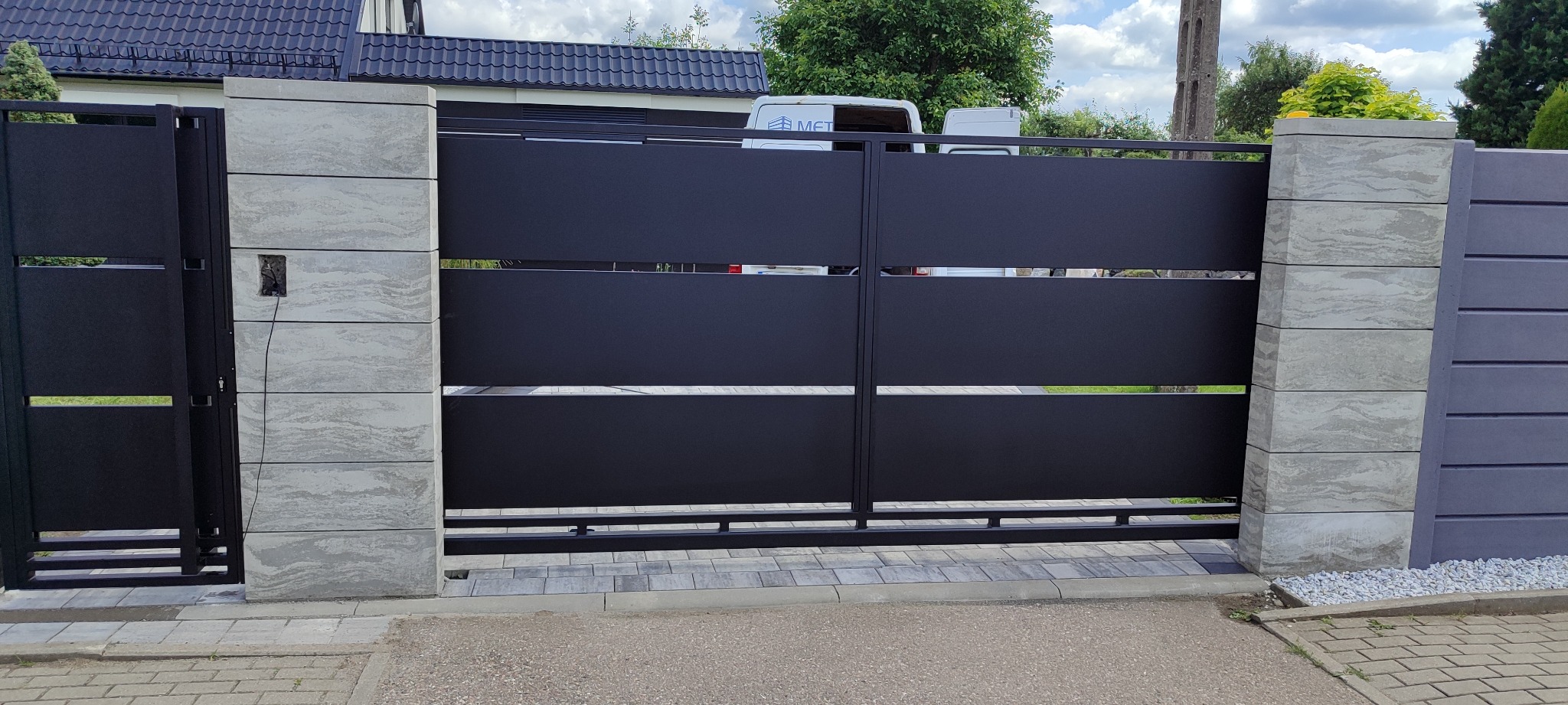 Nowoczesna brama wjazdowa z czarnymi, poziomymi panelami i betonowymi słupkami z widoczną instalacją domofonową po lewej stronie. Tło: dom z ciemną dachówką.