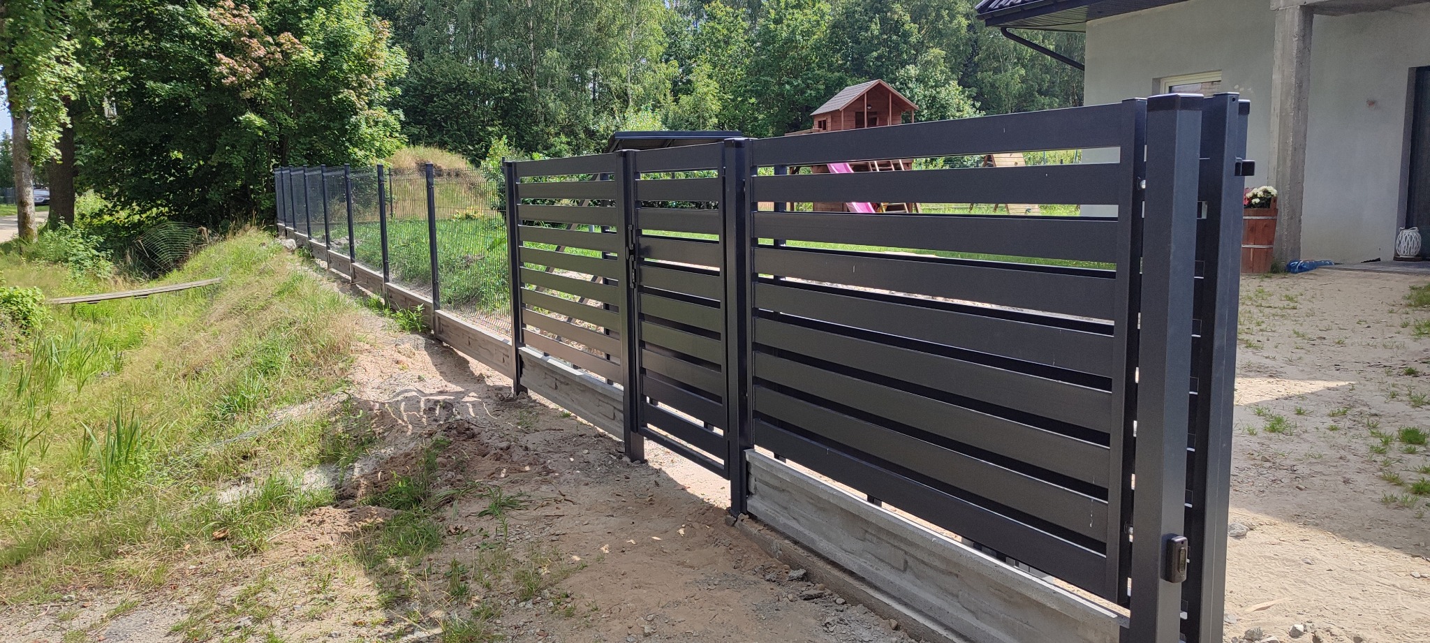 Nowoczesne, poziome ogrodzenie panelowe w kolorze antracytowym, z betonową podmurówką, na tle zieleni i domu. Widok z zewnątrz posesji, w słoneczny dzień.