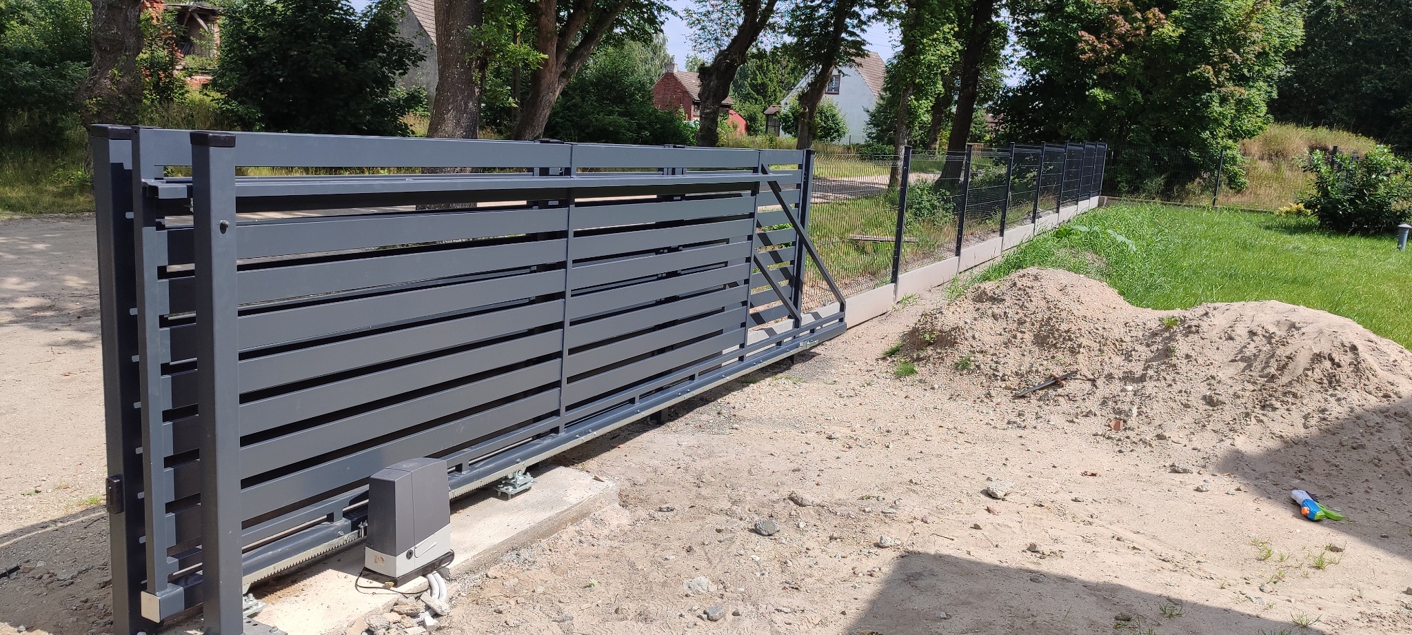 Nowoczesna, przesuwna brama wjazdowa w kolorze grafitowym, z widocznym napędem, zamontowana przy ogrodzeniu panelowym na tle zieleni.