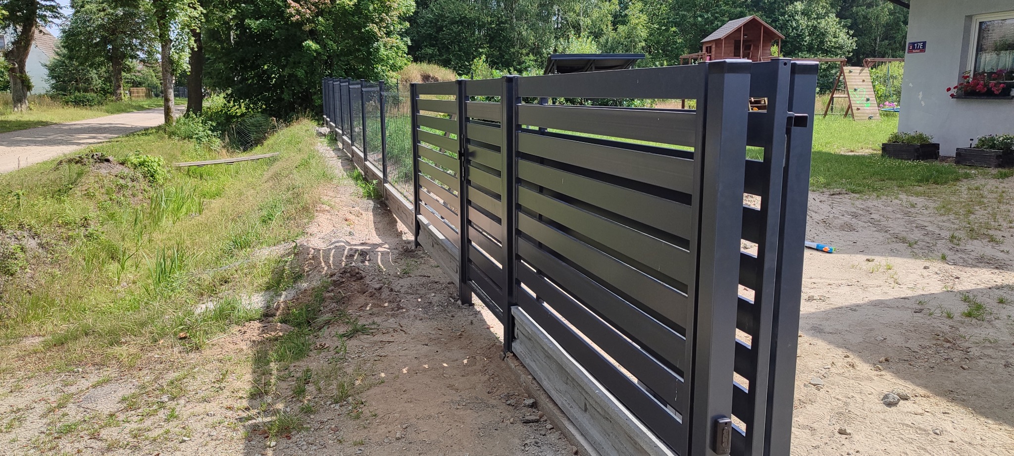 Nowoczesna, antracytowa brama wjazdowa z poziomymi panelami, zintegrowana z ogrodzeniem panelowym. Widok z zewnątrz posesji, w tle dom z placem zabaw.