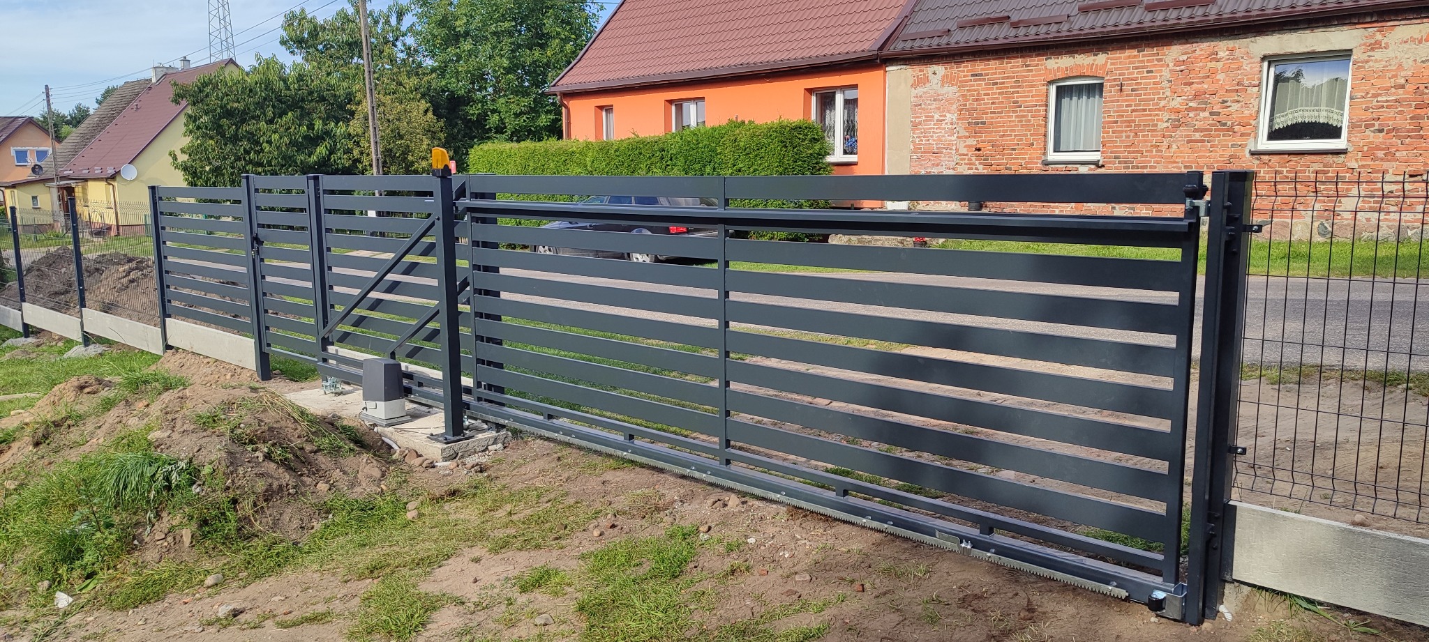 Nowoczesna, przesuwna brama wjazdowa z poziomymi panelami w kolorze grafitowym, z widocznym napędem, na tle domu z czerwonej cegły i zieleni.