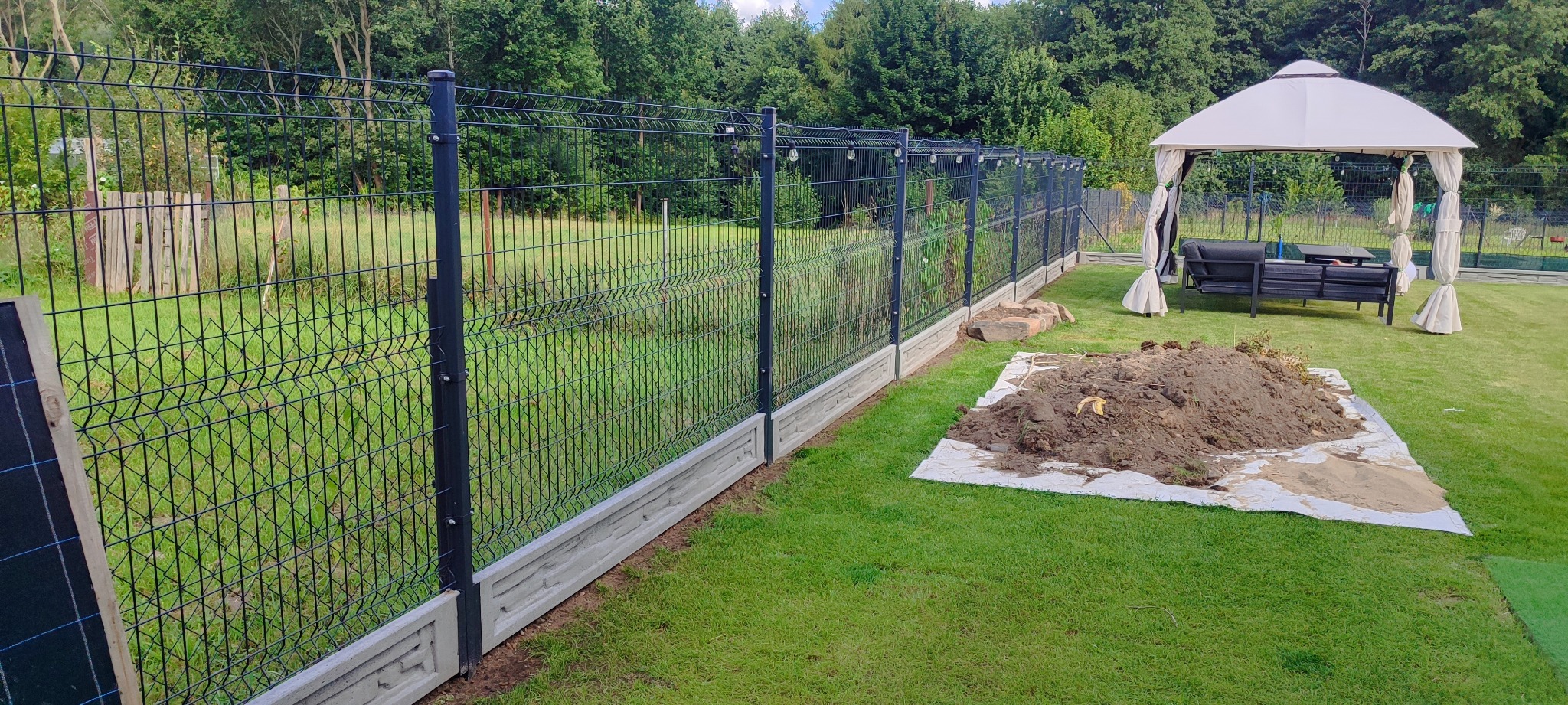 Nowoczesne ogrodzenie panelowe z podmurówką betonową na zielonej działce, w tle altana ogrodowa. Widok na zainstalowane ogrodzenie w słoneczny dzień.