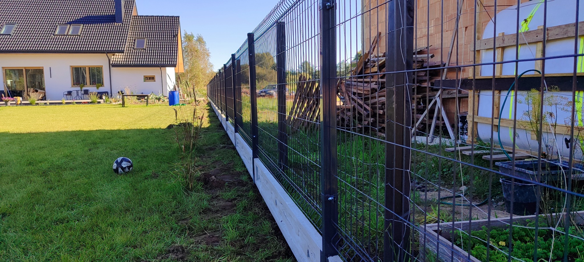 Nowoczesne ogrodzenie panelowe z podmurówką betonową na tle domu jednorodzinnego i zielonego trawnika z piłką. Widoczne elementy konstrukcyjne i materiały budowlane.