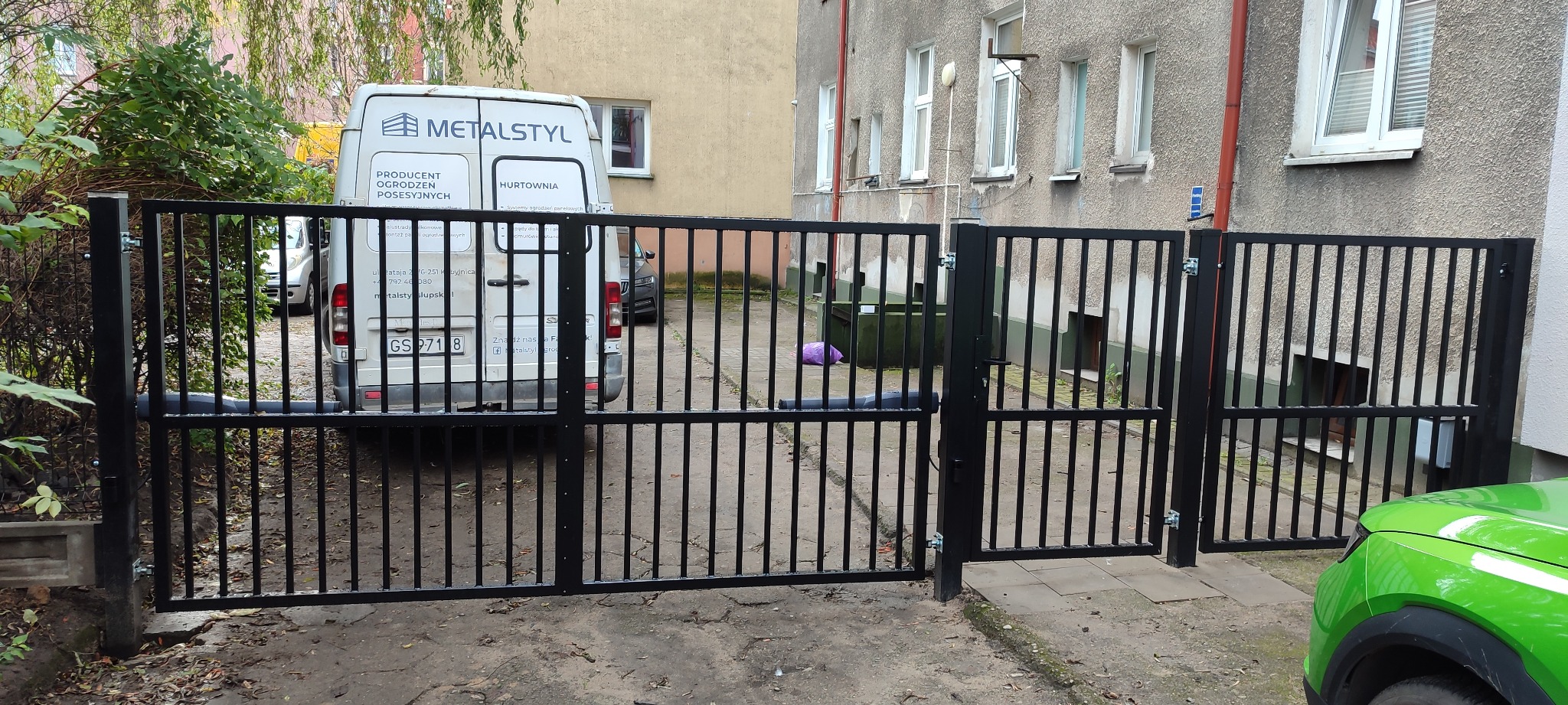 Czarna, metalowa brama wjazdowa na posesję w Kobylnicy, widoczna z bliska. W tle budynek i samochód dostawczy z logo firmy.