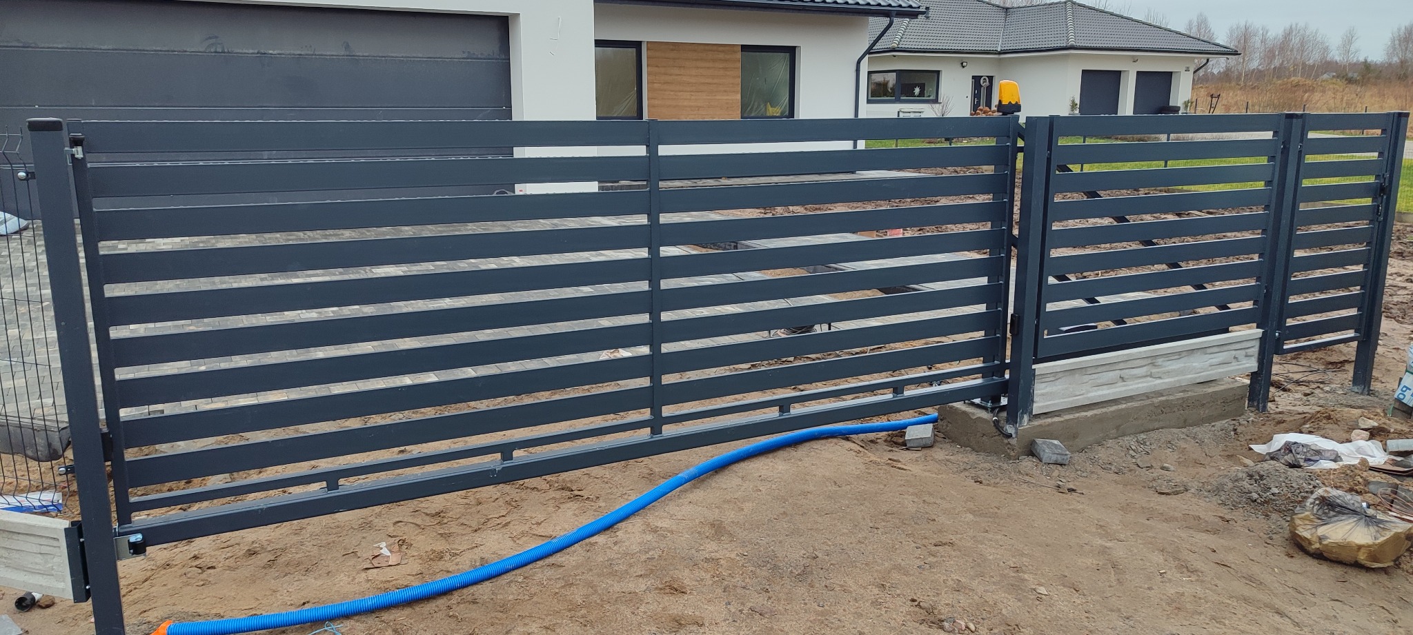 Nowoczesna, pozioma brama przesuwna z szarego metalu, z widocznym napędem i fragmentem ogrodzenia panelowego. W tle dom jednorodzinny.