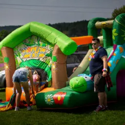 Rozłożony, dmuchany plac zabaw z motywem dżungli na trawie, w tle osoba w hawajskim naszyjniku i okulary przeciwsłoneczne oraz sylwetka dziecka wchodzącego do środka.