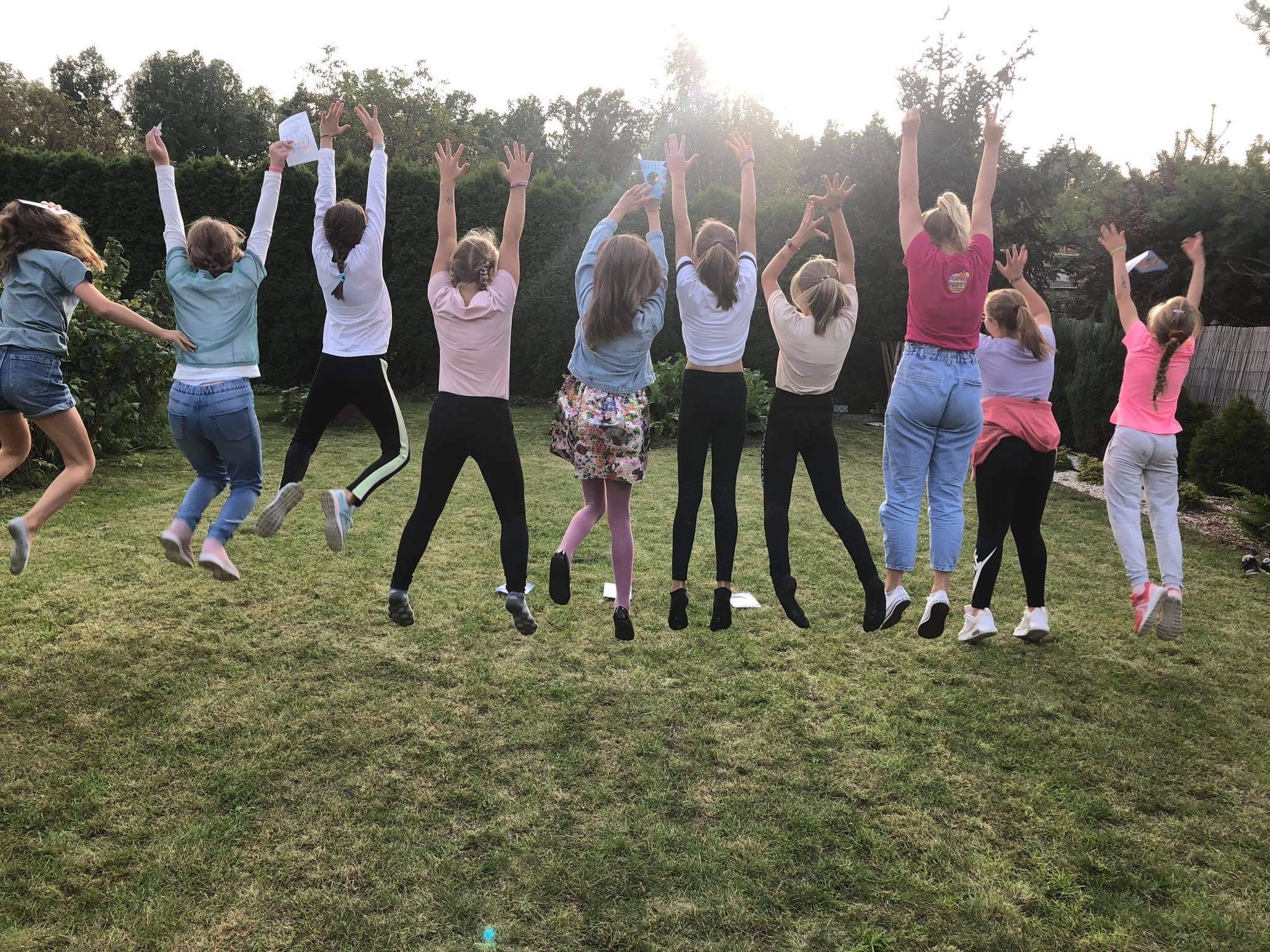 Grupa dziewcząt w różnym wieku skacze na trawie z podniesionymi rękami, trzymając kartki; w tle zielone krzewy i drzewa.