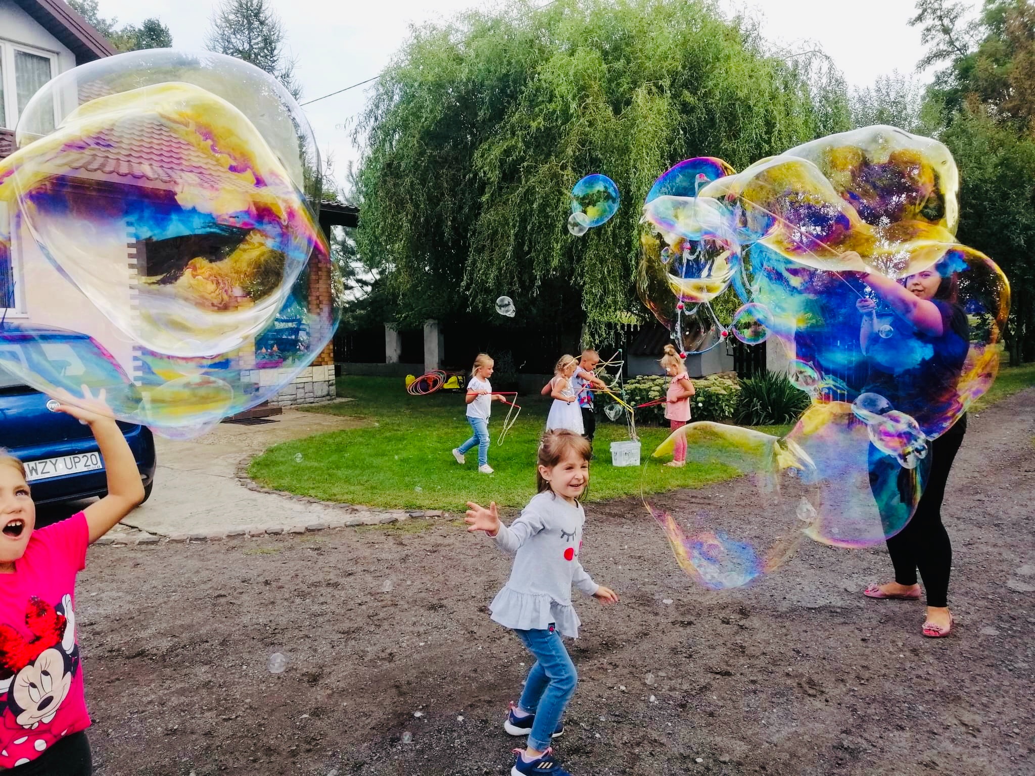 Dziewczynka w różowej koszulce z Myszką Minnie łapie ogromną bańkę mydlaną, w tle inne dzieci i osoba tworząca bańki o różnych rozmiarach.