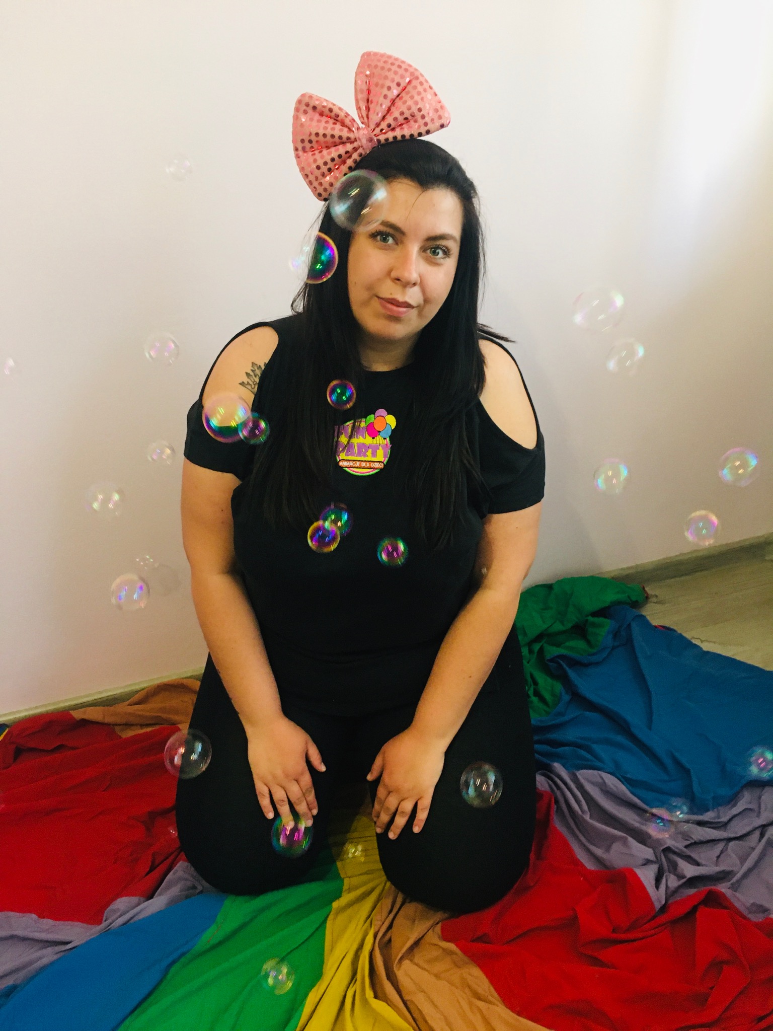 Kobieta w czarnym stroju, z kokardą na głowie, klęczy na kolorowej tkaninie, dmuchając bańki mydlane w pomieszczeniu z białymi ścianami.