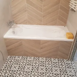 Nowa wanna zabezpieczona folią, otoczona płytkami ułożonymi w jodełkę i wzorzystą posadzką, widoczne elementy instalacji wodnej i grzejnik drabinkowy.