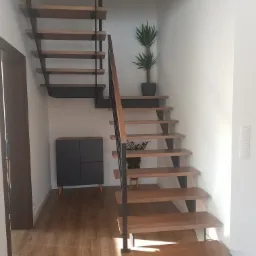 Drewniane schody ażurowe z czarnymi metalowymi wspornikami w nowoczesnym wnętrzu z drewnianą podłogą, szafką i roślinami doniczkowymi.