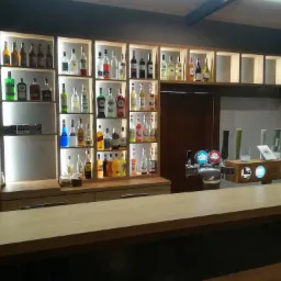 Drewniane regały z podświetleniem, wypełnione butelkami alkoholi, nad blatem baru w Jawiszowicach. Widoczne dystrybutory piwa.