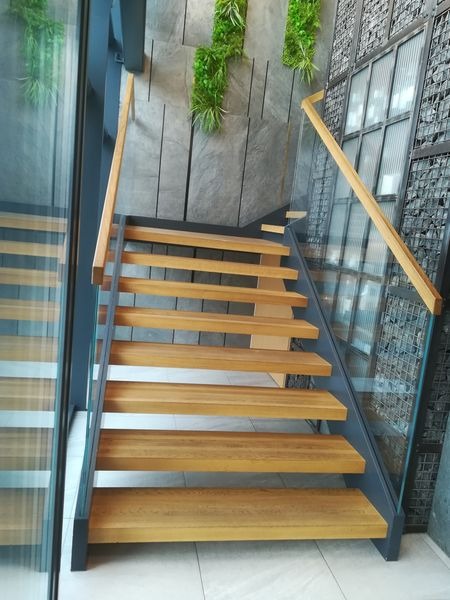 Nowoczesne schody drewniane z metalową konstrukcją i szklaną balustradą, oświetlone naturalnym światłem, z dekoracyjną ścianą z zielenią w tle.