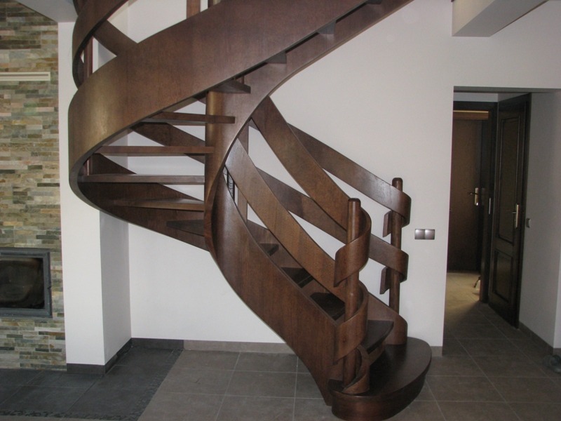 Drewniane schody spiralne o ciemnym odcieniu brązu, z dekoracyjnymi, zakręconymi elementami poręczy, umieszczone w przestronnym wnętrzu z kamienną ścianą kominkową w tle.