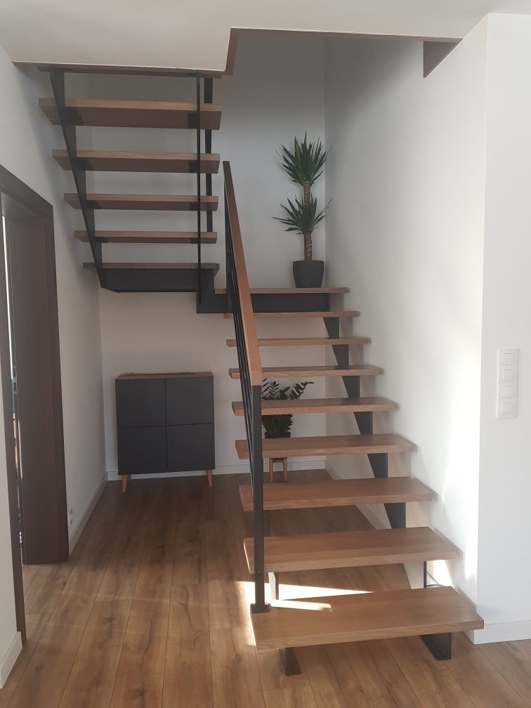 Drewniane schody ażurowe z czarnymi metalowymi wspornikami w nowoczesnym wnętrzu z drewnianą podłogą, szafką i roślinami doniczkowymi.