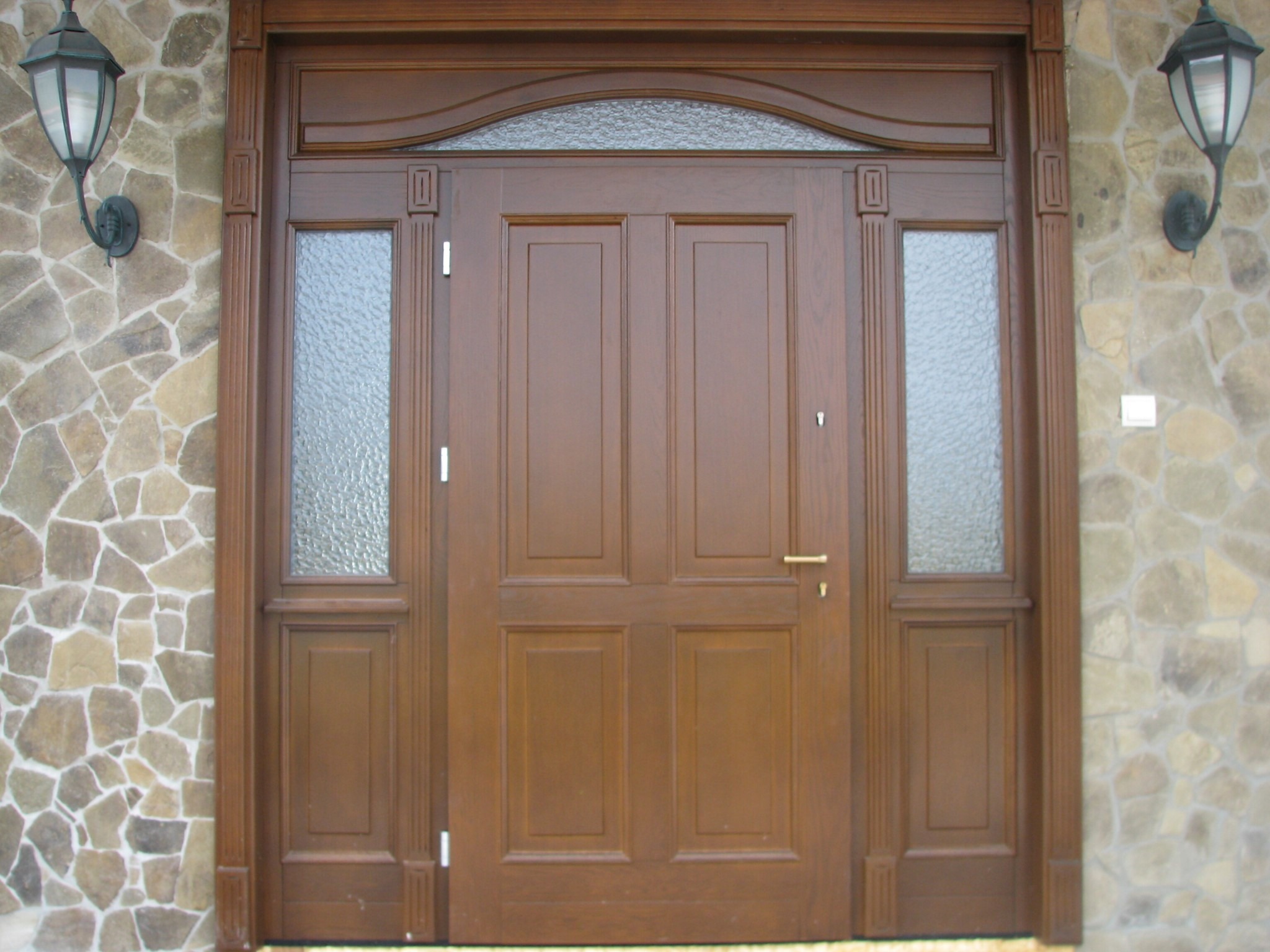 Solidne, dwuskrzydłowe drzwi wejściowe z drewna w kolorze orzechowym, z bocznymi szybami o fakturze lodu i górnym łukowym świetlikiem, osadzone w kamiennej elewacji z dwoma lampami ściennymi.